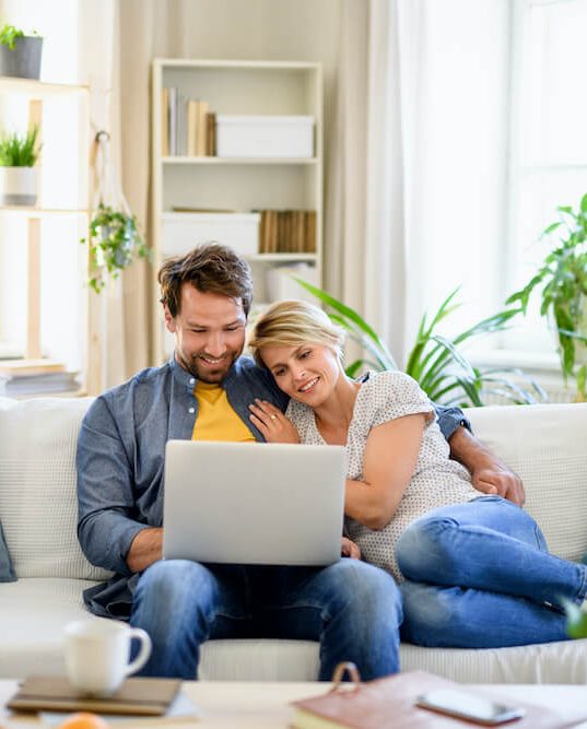 Stock-style image of a couple using a laptop while seated together on a sofa in a bright home interior.