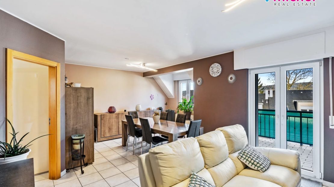 Open dining and living area with a wooden table, cream sofa, storage units, and double glazed doors opening onto a balcony.