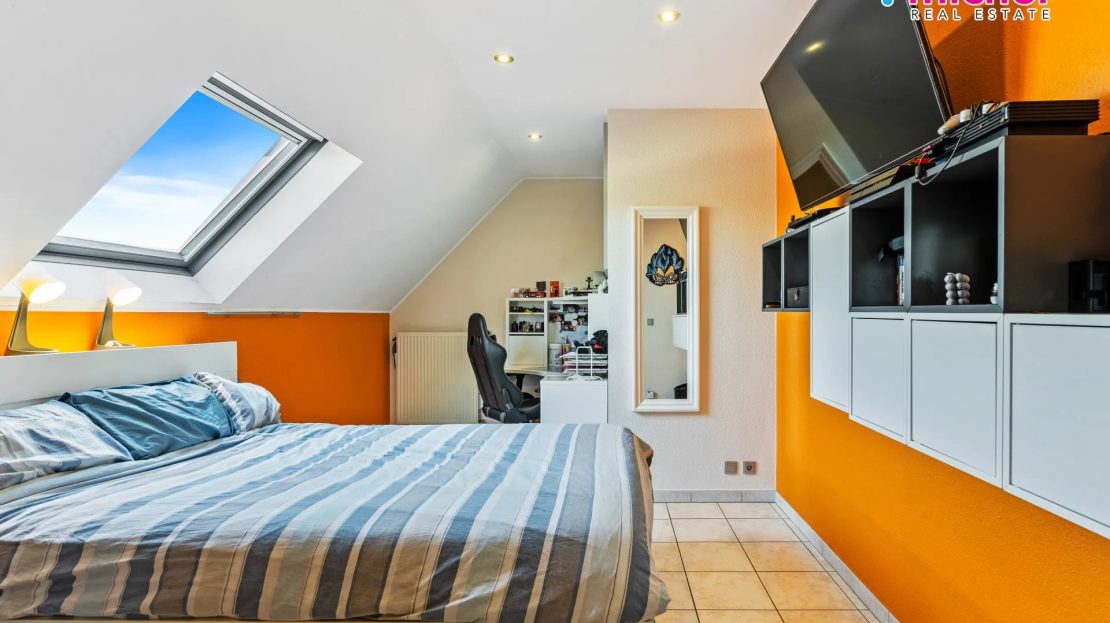 Attic-style bedroom with a sloped ceiling, skylight, orange accent wall, bed, desk, mirror, and wall-mounted television.