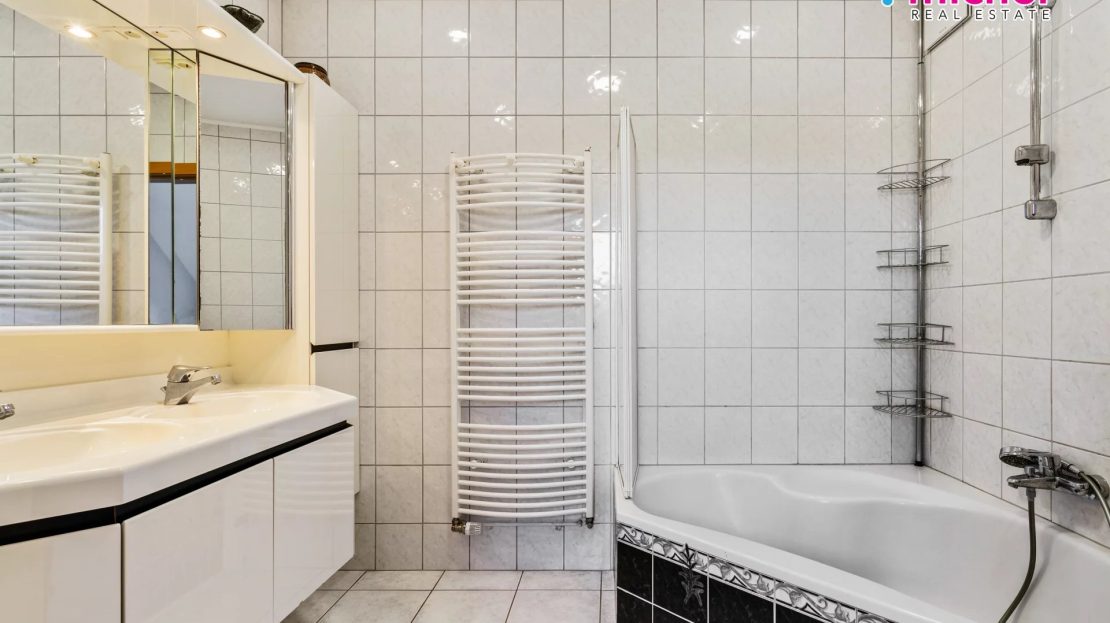 Bright tiled bathroom with a bathtub, shower fittings, sink, mirror, and heated towel rail.